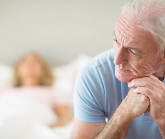 Oudere man zit aan de rand van het bed en kijkt met bedenkelijke blik uit het raam.