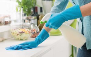 Vrouw met blauwe handschoenen maakt keukenaanrecht schoon met schoonmaakspray en doekje.