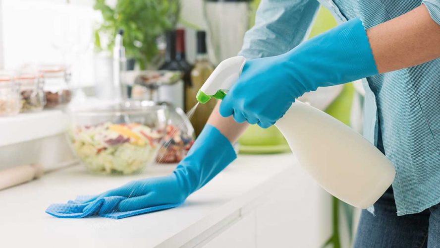 Vrouw met blauwe handschoenen maakt keukenaanrecht schoon met schoonmaakspray en doekje.