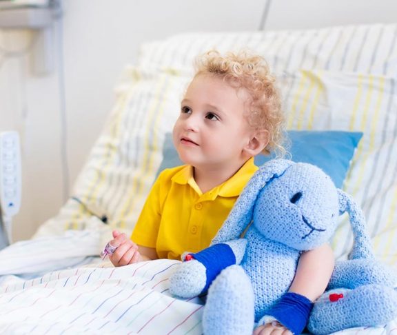 Jongen met blonde krullen op ziekenhuisbed met blauw knuffelkonijn.