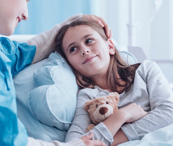 Meisje met knuffelbeer op ziekenhuisbed, glimlacht naar veprleegkundige.